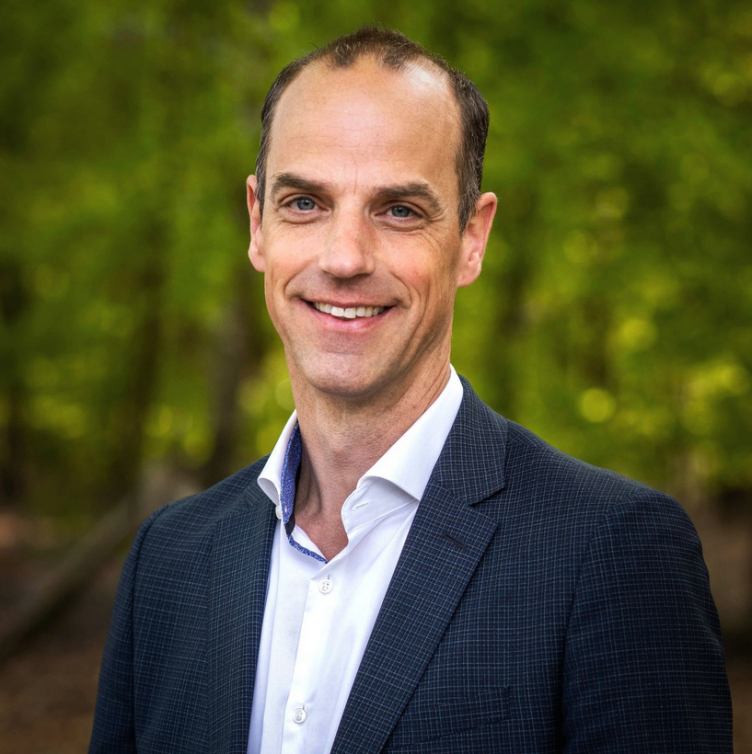 Portrait of a tall, balding caucasian man in dressshirt and jacket. He is smiling and looking straight at the camera. Blurry trees in the background.