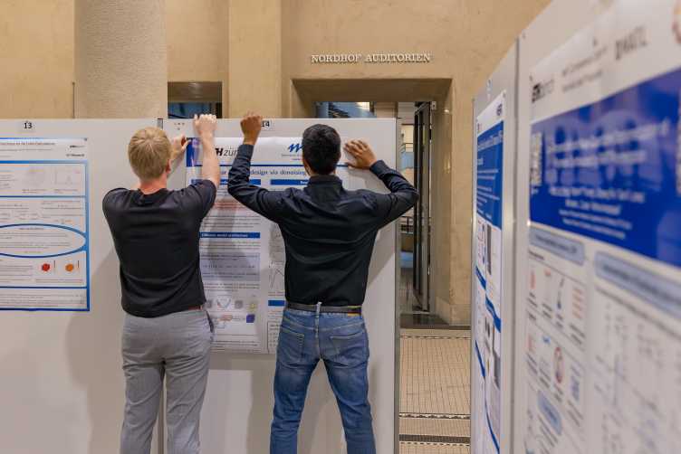 2 guys, seen from behind, hanging up a scientific poster on a poster wall