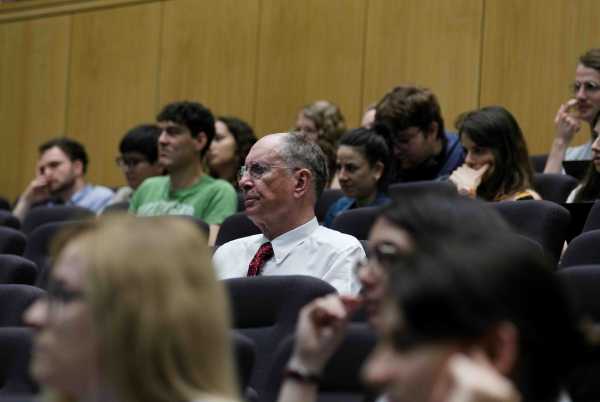 People in a lecture hall, in the middle a caucasian man in his 60s wearing a white shirt with red tie and glasses