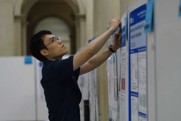 An Asian man with glasses pinning up his poster. Seen from the side.