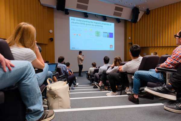 Perspective from above the stairs in a lecture hall. Presenter in front of a screen, audiences to the right and left