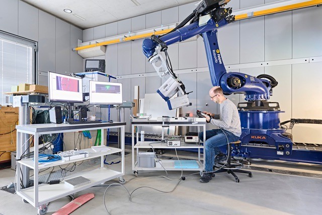 A man sitting next to a shiny dark blue robotic device with a long arm, two computer screens on his left