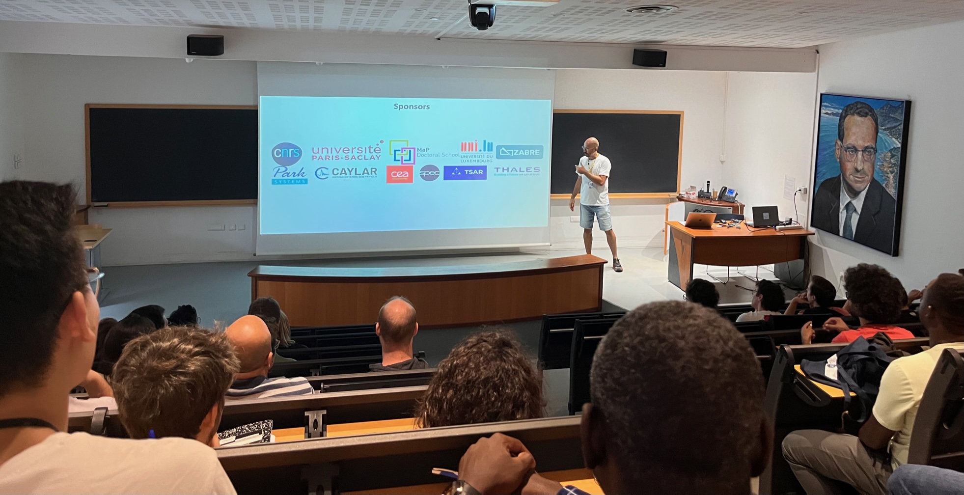 A person holding a lecture in a lecture hall. On the screen several sponsor logos.
