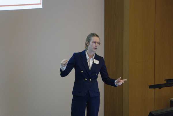 A caucasian woman with glasses and ponytail in a dark suit gesticulating while holding a talk