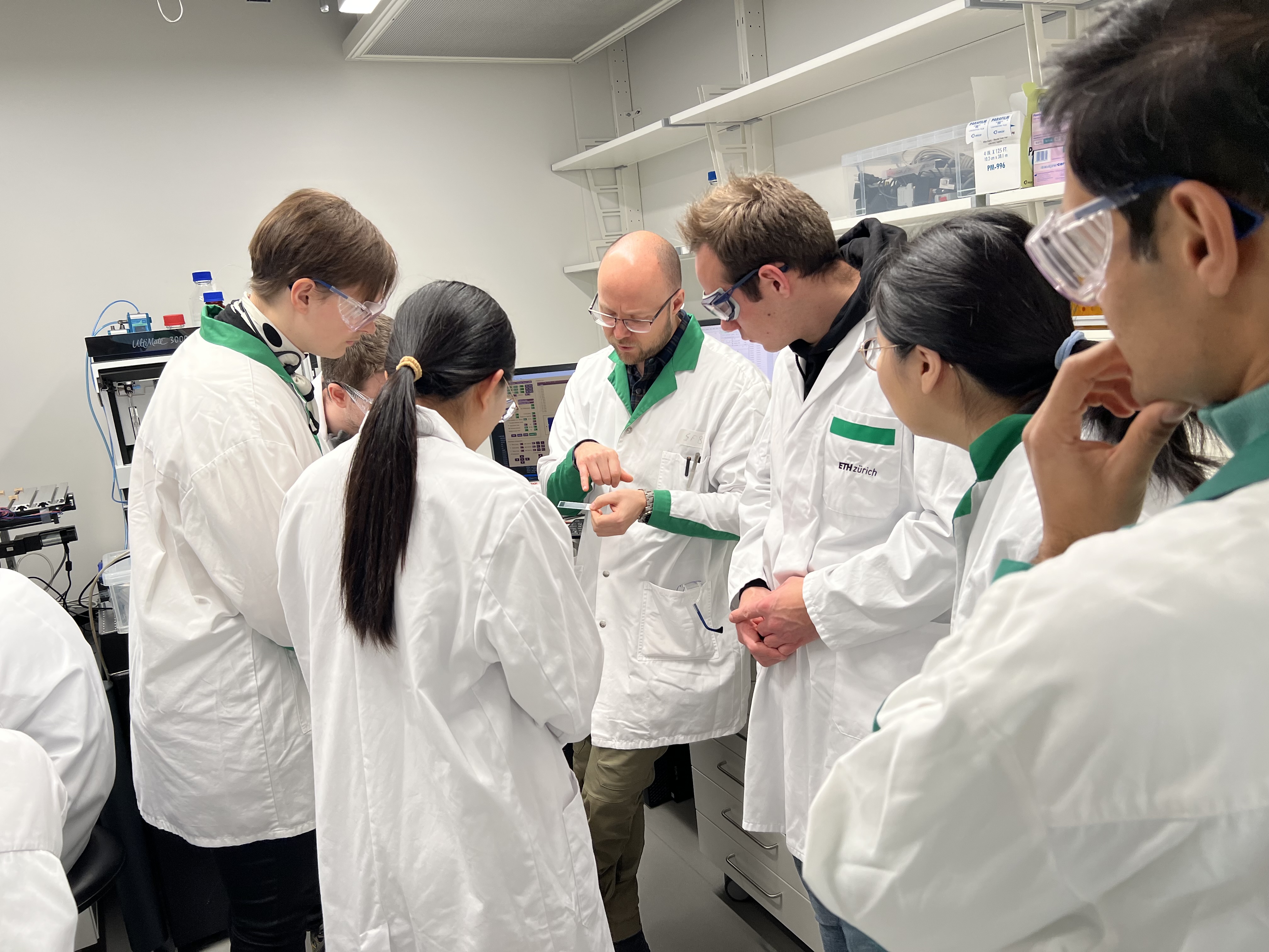 A lab scene. Around 6 people wearing lab coats and protective goggles looking at a glass slide, which one of them points to.