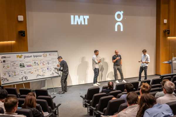 inside Audi Max, three people on stage talking, IMT and ON logos on the screen. Graphic illustrator working on his illustration on the left.