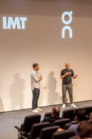 Two guys on stage in Audi Max, ON and IMT logo displayed behind them