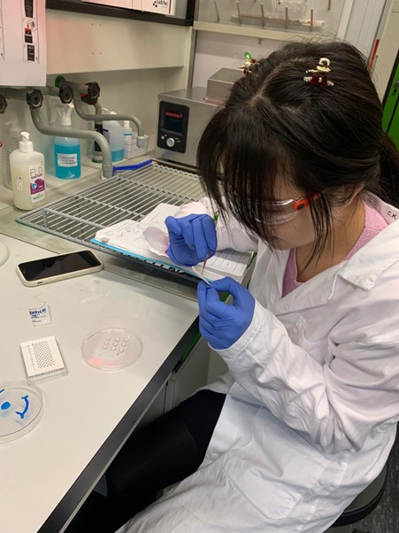 A young Asian woman sitting at a lab bench, wearing a lab coat, goggles, and blue plastic gloves