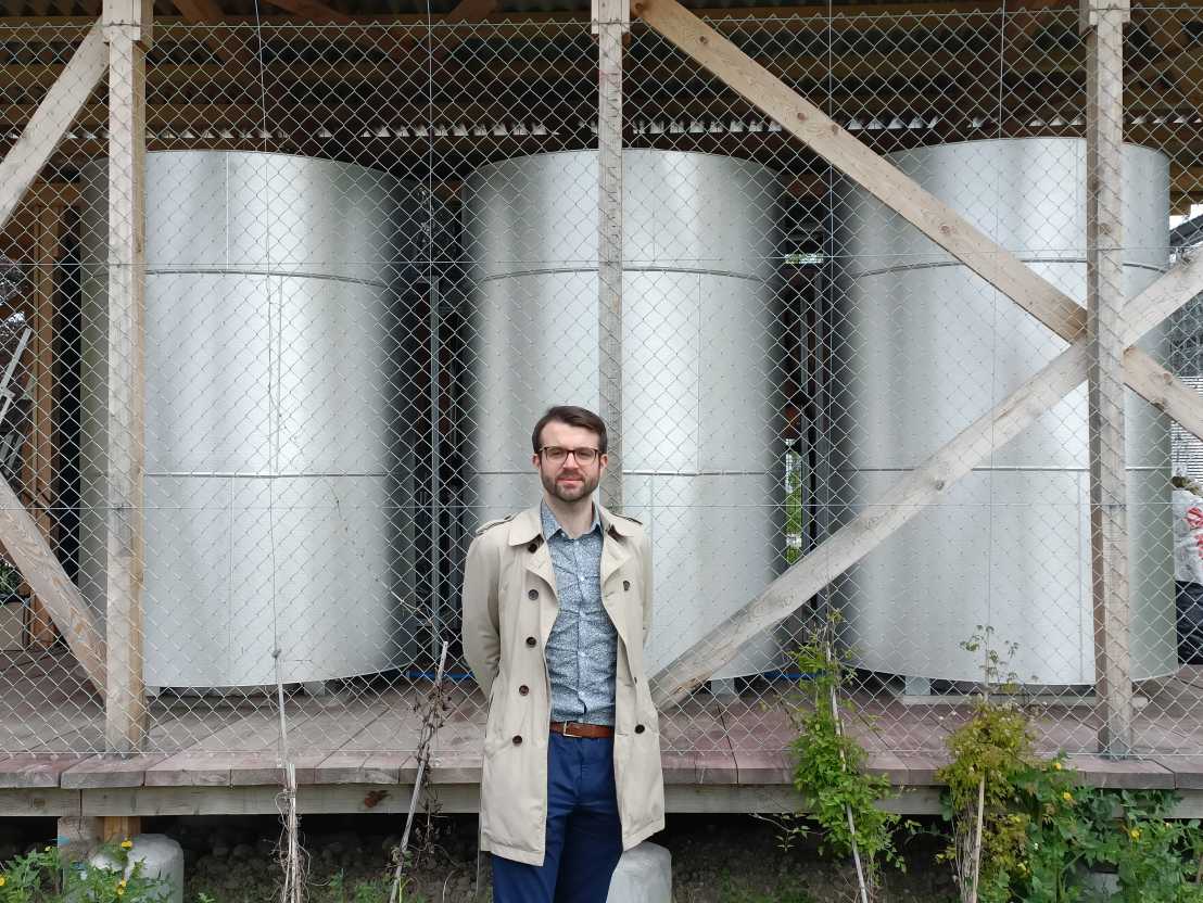 A young man in a light coat in front of three silvery cylindrical containers