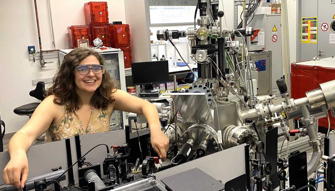 A young woman sitting in a lab with lots of machinery. She is wearing protective goggles and a summer dress.